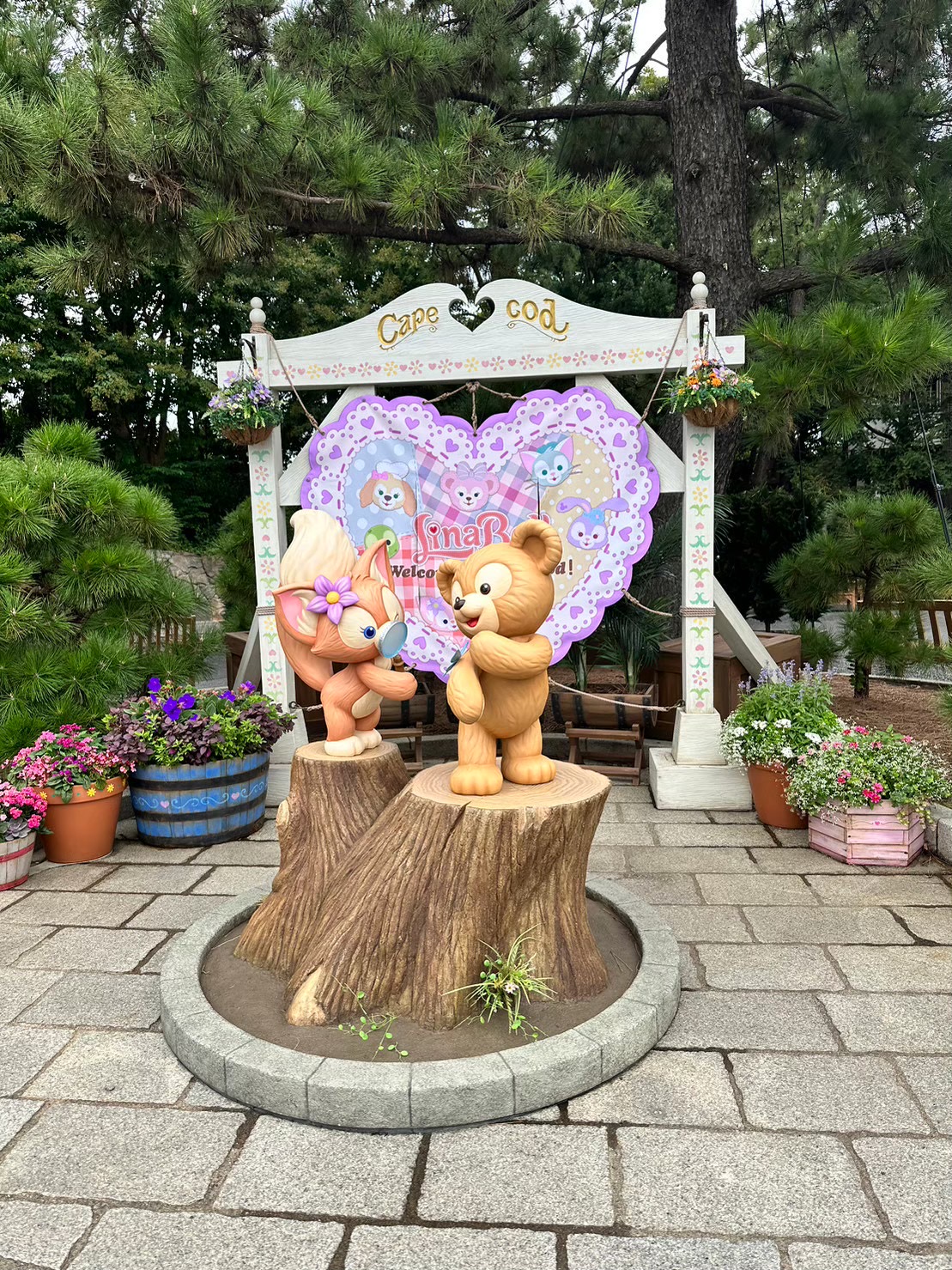 ケープコッドの木の切り株の上に立つダッフィーとリナベルの像。背後にはハート型の看板と花の鉢植えが飾られている。