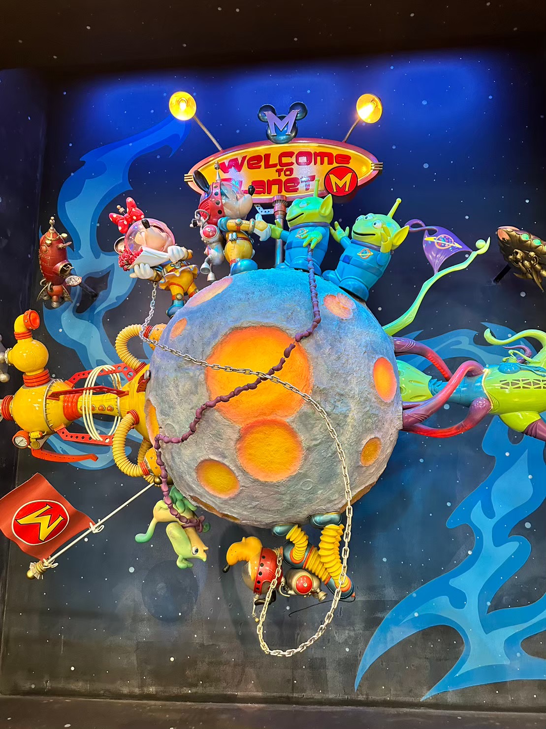 「WELCOME TO PLANET M」の看板の下、宇宙服姿のミニーマウスやトイ・ストーリーのエイリアンたちが月面オブジェに集まっている装飾。